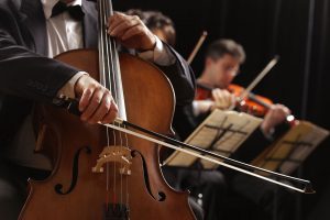 Músico tocando violoncelo. Al fondo una joven tocando el violín con atril