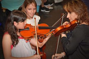 Niña y Profesora tocando el Violín. Otra niña las observa en segundo plano