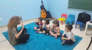 Niños sentados jugando con profesora guitarra al fondo