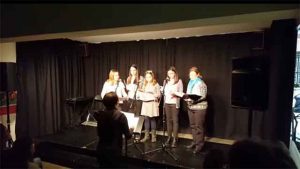 Coro de Alumnos cantando sobre escenario. Profesora de espalda dirigiendo